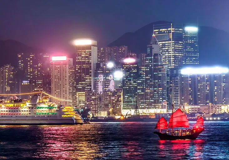 Hong Kong harbor at night with illuminated buildings and a red-sailed boat