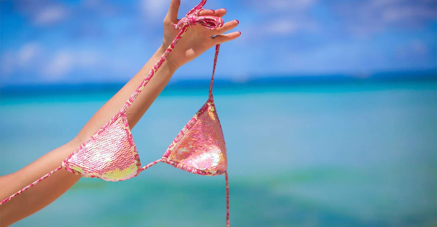 Una mano sosteniendo un top de bikini rosa contra un mar turquesa