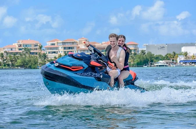 Two riders on a jet ski with Cancun waterfront hotels behind them